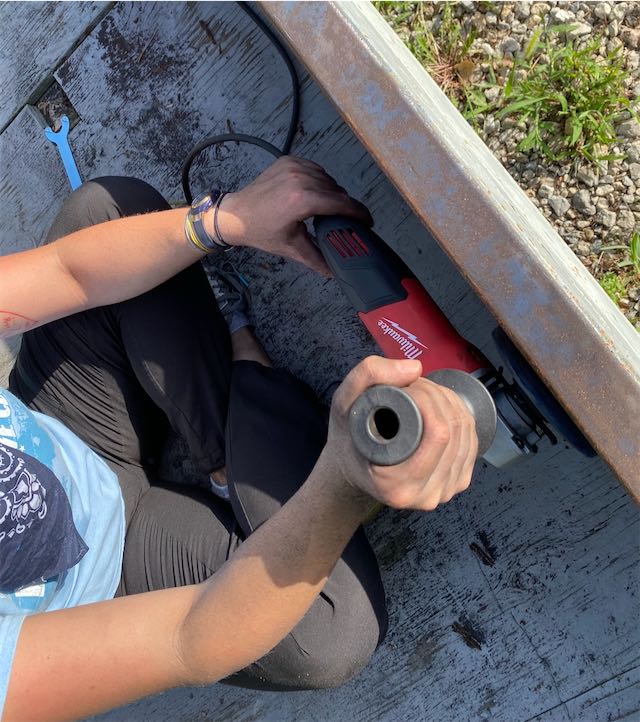 Shows a young woman using an angle grinder to remove rust from metal.